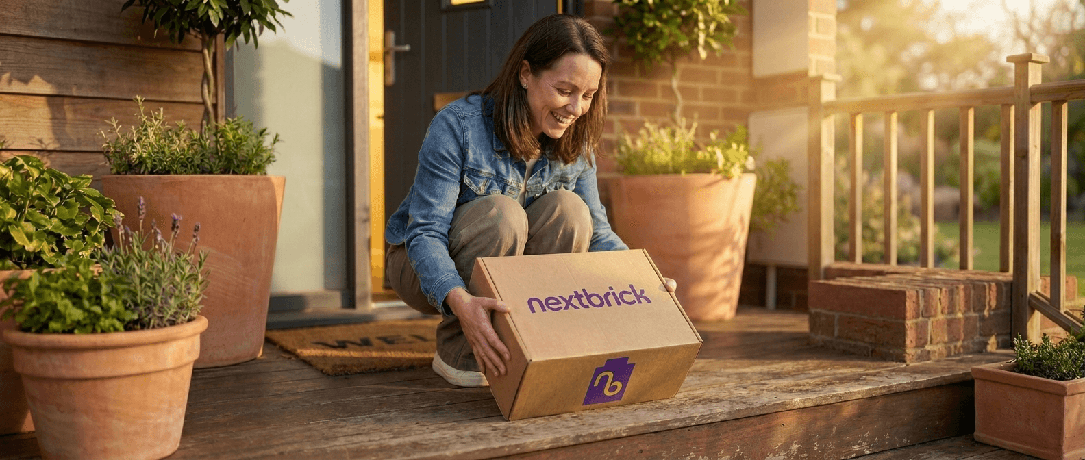 A smiling woman on a front porch picking up a Next Brick package, with the front door open behind her in warm golden-hour light